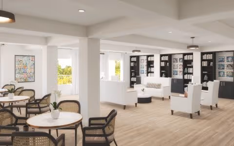 A bright and spacious common area with light wood flooring, white walls, and large windows letting in natural light. The room is furnished with round tables and cane-backed chairs on the left side, and white cushioned armchairs arranged around a small table on the right. Built-in black bookshelves filled with books and decorative items line the back wall. Modern ceiling lights hang from the ceiling.