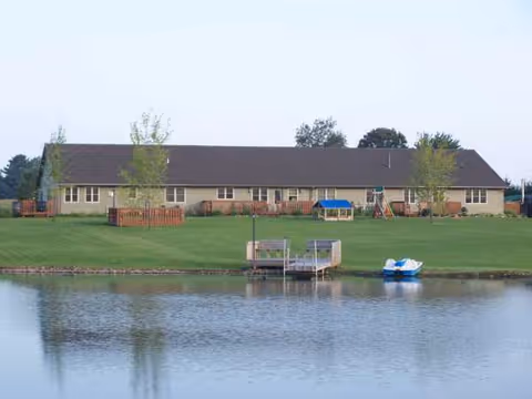 A single-story building sits along a grassy lawn beside a calm lake, with a dock.