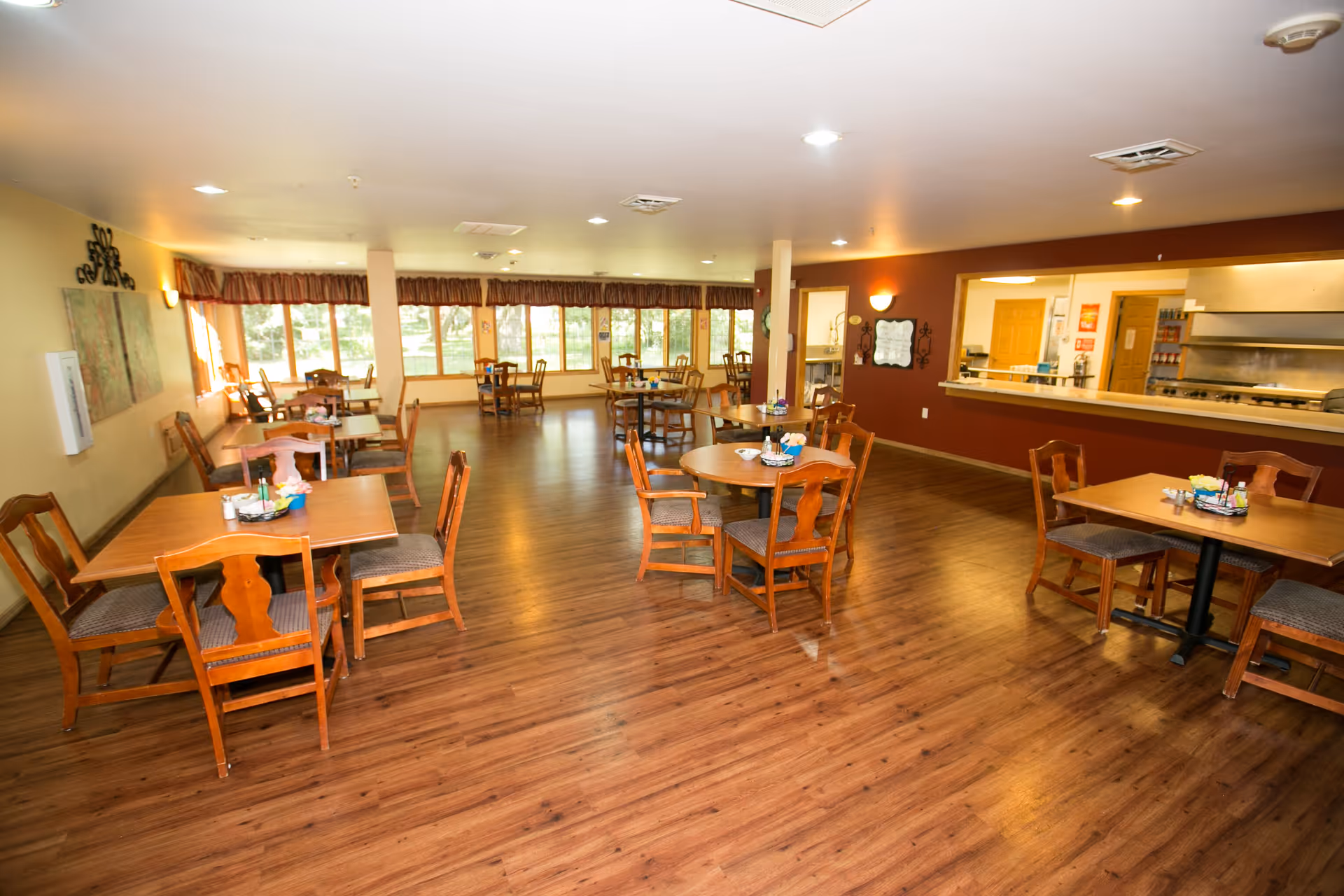 A spacious dining room in a senior living facility with wooden tables and chairs arranged neatly on a wood floor. Large windows along one wall let in natural light, and there is a serving window to the kitchen area with stainless steel appliances visible. The walls are painted in warm tones with some decorative elements.