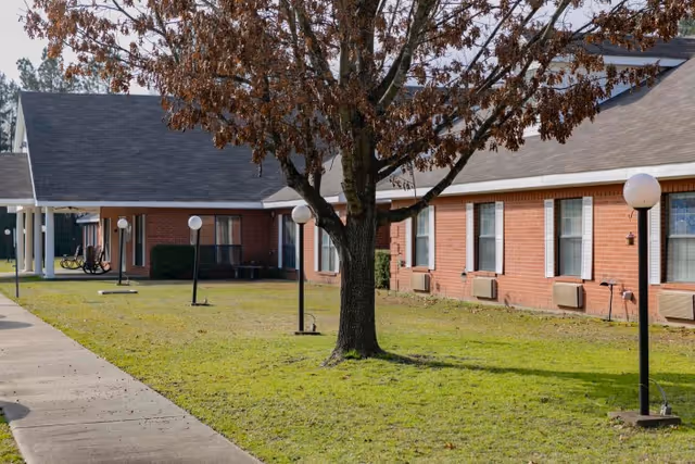 Front exterior of a single-story brick senior care building with a grassy courtyard, a central tree, sidewalk, and lamp posts.