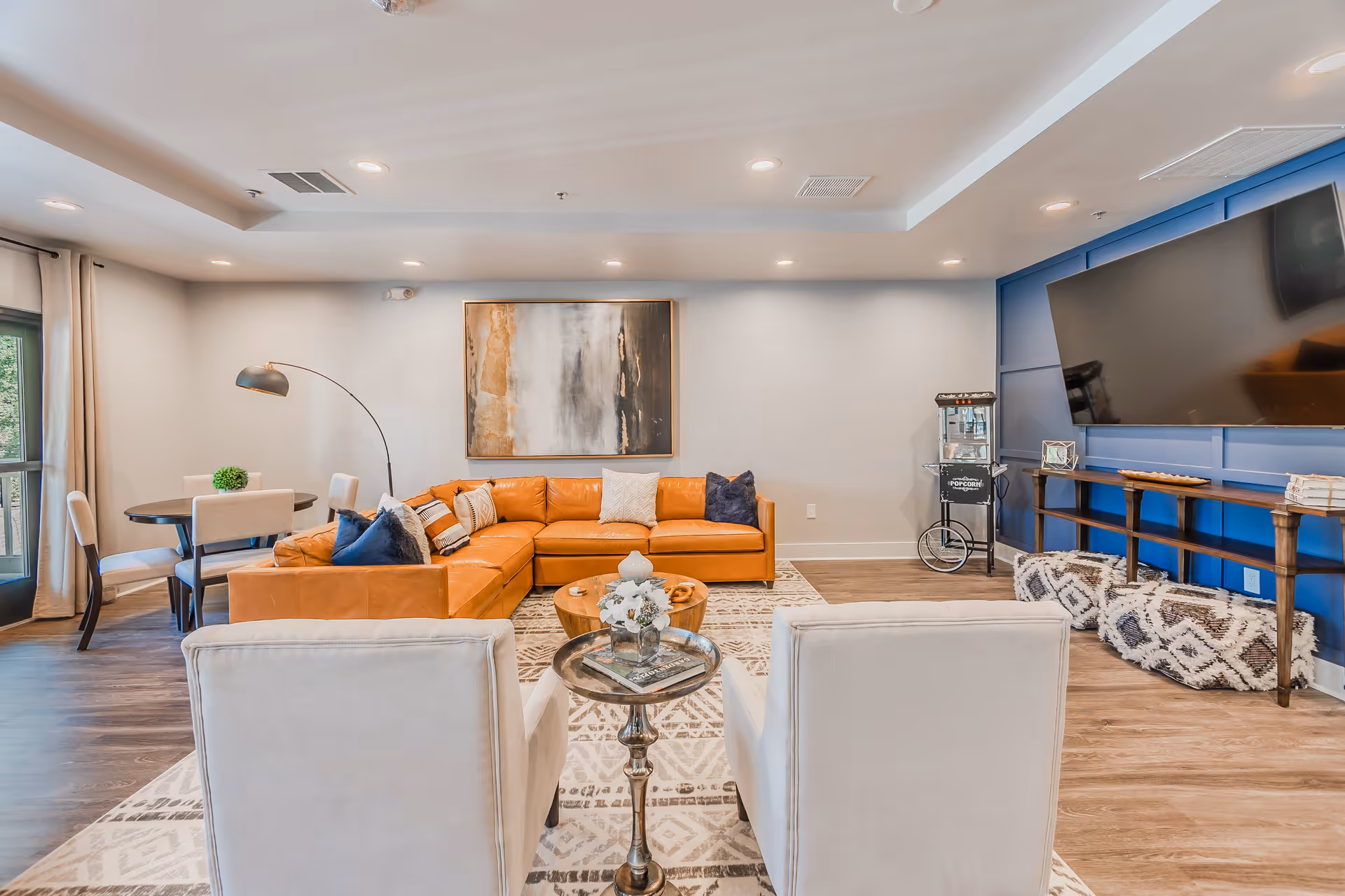 A modern living room with a large orange sectional sofa adorned with various pillows, two white armchairs facing the sofa with a small round table between them, a round wooden coffee table, a large flat-screen TV mounted on a blue accent wall, a popcorn machine on a stand, and a dining table with four chairs near a window with curtains.