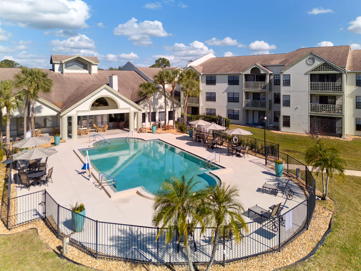 Outdoor swimming pool area at Cascade Heights facility with lounge chairs, tables with umbrellas, palm trees, and a multi-story building in the background under a partly cloudy blue sky.