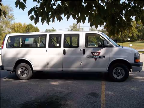 White Sweet Springs Villa van parked in a lot under tree shade, logo visible