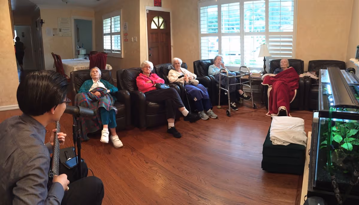 A group of elderly people sitting in a living room on leather chairs arranged in a semi-circle, listening to a man playing a guitar and singing into a microphone. The room has wooden floors, large windows with white shutters, and a fish tank on the right side.