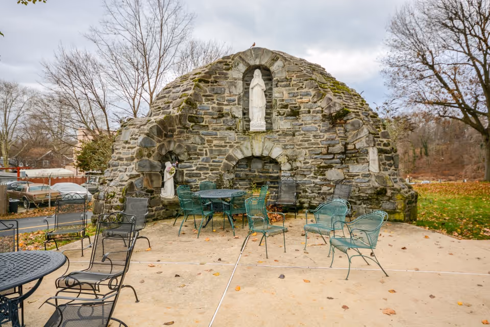 Outdoor seating area with metal chairs and tables arranged on a concrete patio in front of a large stone grotto featuring religious statues. Trees without leaves and a cloudy sky are visible in the background.