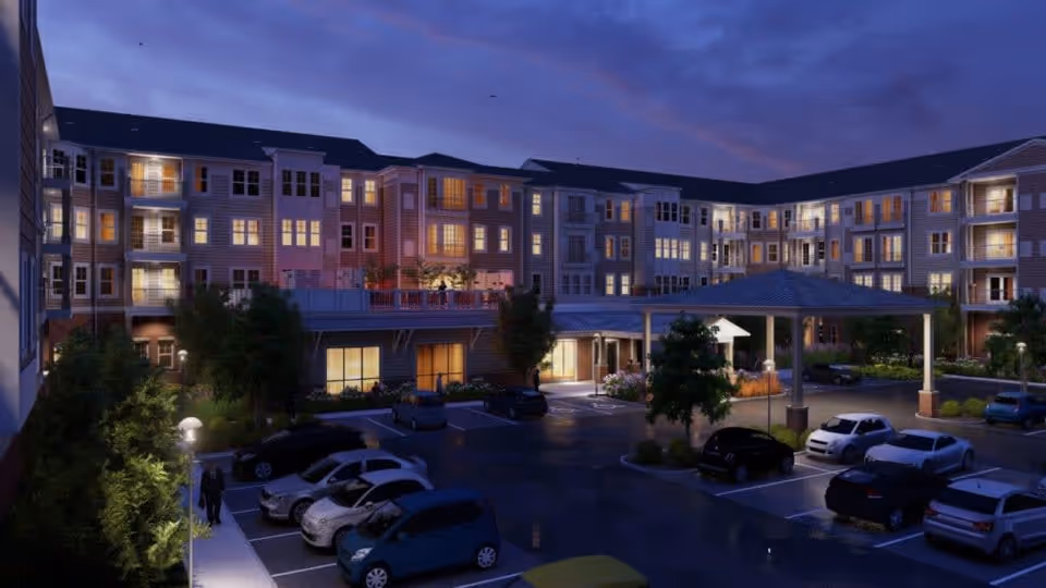Evening view of a multi-story senior living facility named Acclaim At East Beach with illuminated windows and a covered entrance. Several cars are parked in the parking lot in front of the building, and trees and shrubs are visible around the area.