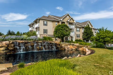 A multi-story residential building with beige siding and white trim, surrounded by green trees and landscaping. In the foreground, there is a small pond with a cascading waterfall made of rocks. The sky is blue with some clouds.