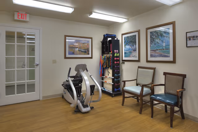A small exercise room with a recumbent exercise bike, a rack holding various exercise equipment including dumbbells and resistance bands, two wooden chairs with cushions, and framed artwork on the walls. There is a white door with glass panels and an exit sign above it.
