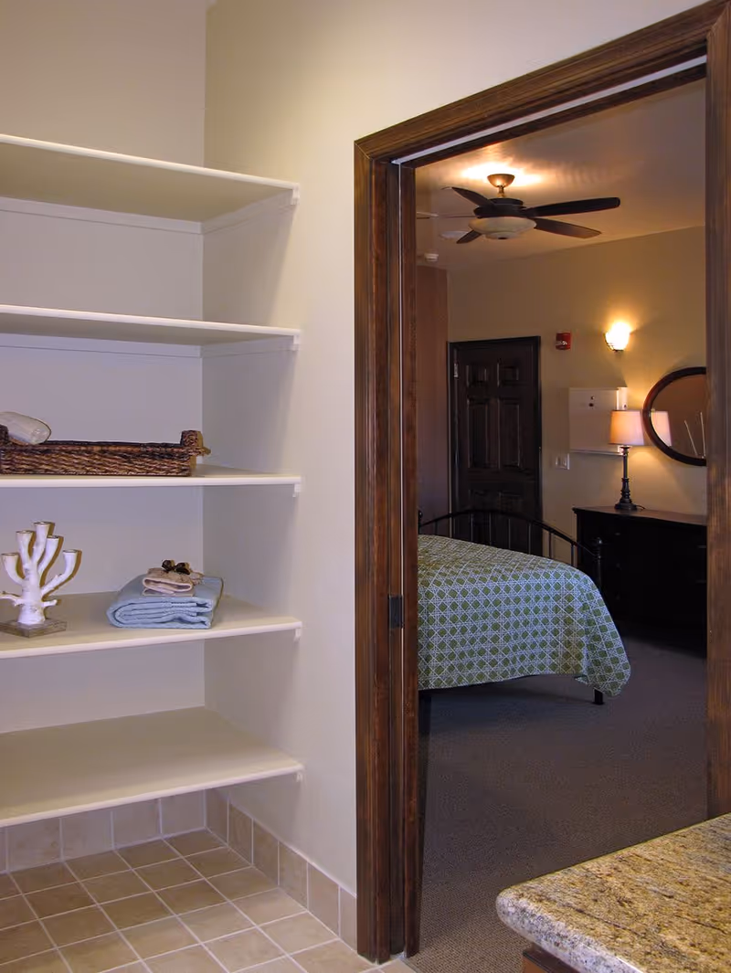 View of a small storage area with white shelves holding a wicker basket, folded towels, and a decorative item, adjacent to a doorway leading into a bedroom with a bed covered in a green patterned bedspread, a ceiling fan, a lamp on a dresser, and a round mirror on the wall.
