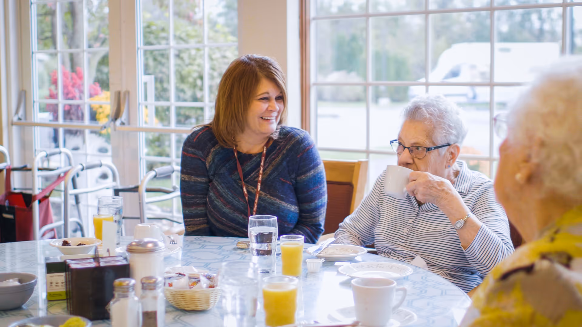 Three people—two older adults and a woman—sitting at a round table with cups and glasses in a bright communal dining area.