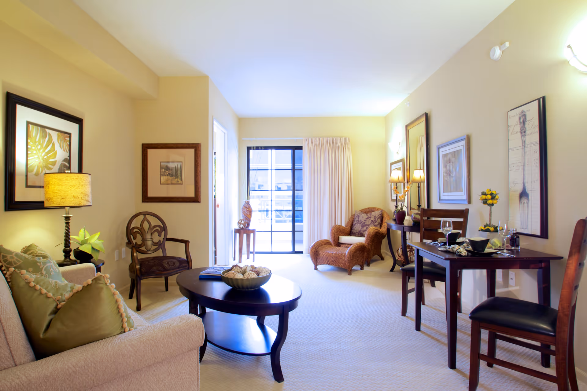 A cozy living room area with a beige sofa adorned with green pillows, a wooden coffee table with decorative items, a wooden chair with a patterned cushion, and a wicker armchair with a cushion. The room has framed artwork on the walls, a table lamp, and a small dining table set with two chairs, dishes, and glasses. A sliding glass door with curtains lets in natural light.