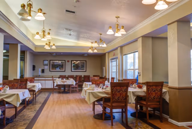 A dining room with several tables covered in white tablecloths and set with plates, glasses, and napkins. The room has wooden chairs with patterned upholstery, beige walls, and multiple ceiling light fixtures. Large windows on one side let in natural light.
