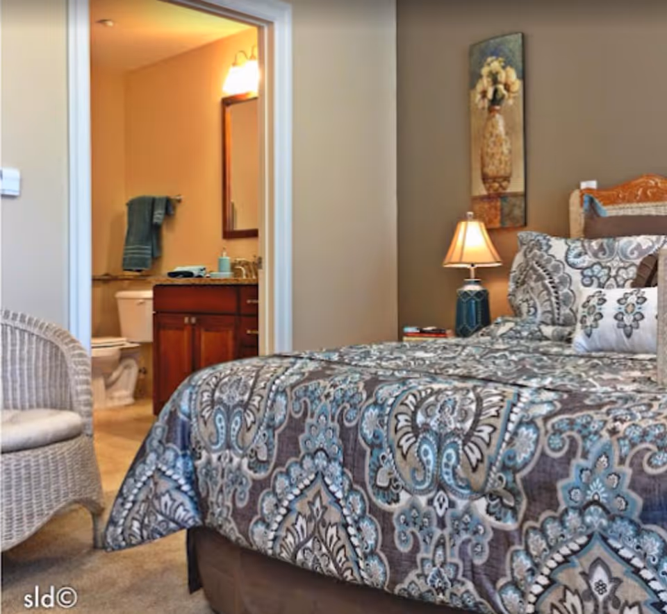 A bedroom with a patterned bedspread in shades of blue, gray, and white. There is a wicker chair with a cushion on the left side. A bedside table with a lamp and some books is next to the bed. On the wall above the bedside table is a decorative painting of a vase with flowers. Through an open door, a bathroom is visible with a toilet, a wooden vanity with a sink, a mirror, and a towel hanging on the wall.