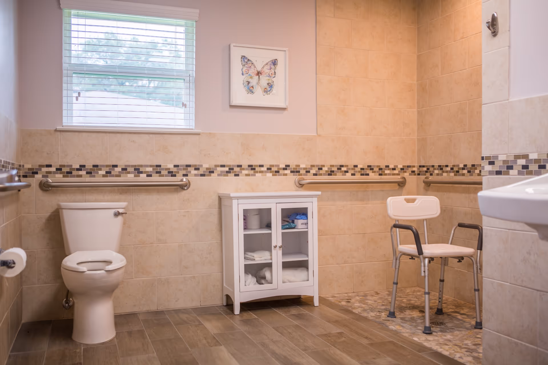 A spacious bathroom with beige tiled walls and wood-look tile flooring. There is a white toilet with a grab bar on the wall beside it, a small white cabinet with glass doors containing towels and supplies, and a walk-in shower area with a white shower chair and grab bars. A window with blinds and a framed butterfly picture are above the cabinet.