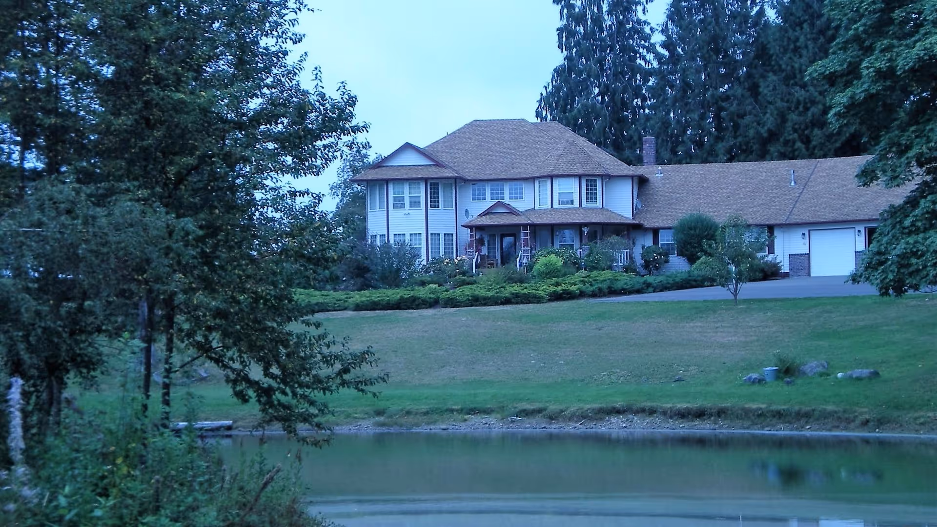 A large two-story house with a brown roof and white exterior situated on a grassy hill next to a small pond, surrounded by trees and greenery.