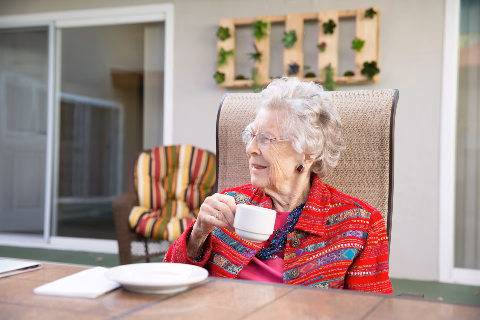 An elderly woman with white hair and glasses sits outdoors in a cushioned chair, holding a white cup and smiling while looking to her left. She is wearing a colorful patterned jacket and a beaded necklace. Behind her is a wall with a decorative wooden planter holding green plants, and a striped cushioned chair is also visible.