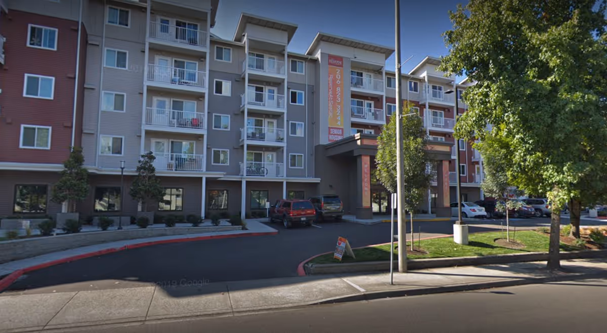 Front exterior of a multi-story residential building with balconies, a covered main entrance and a driveway with parked cars.
