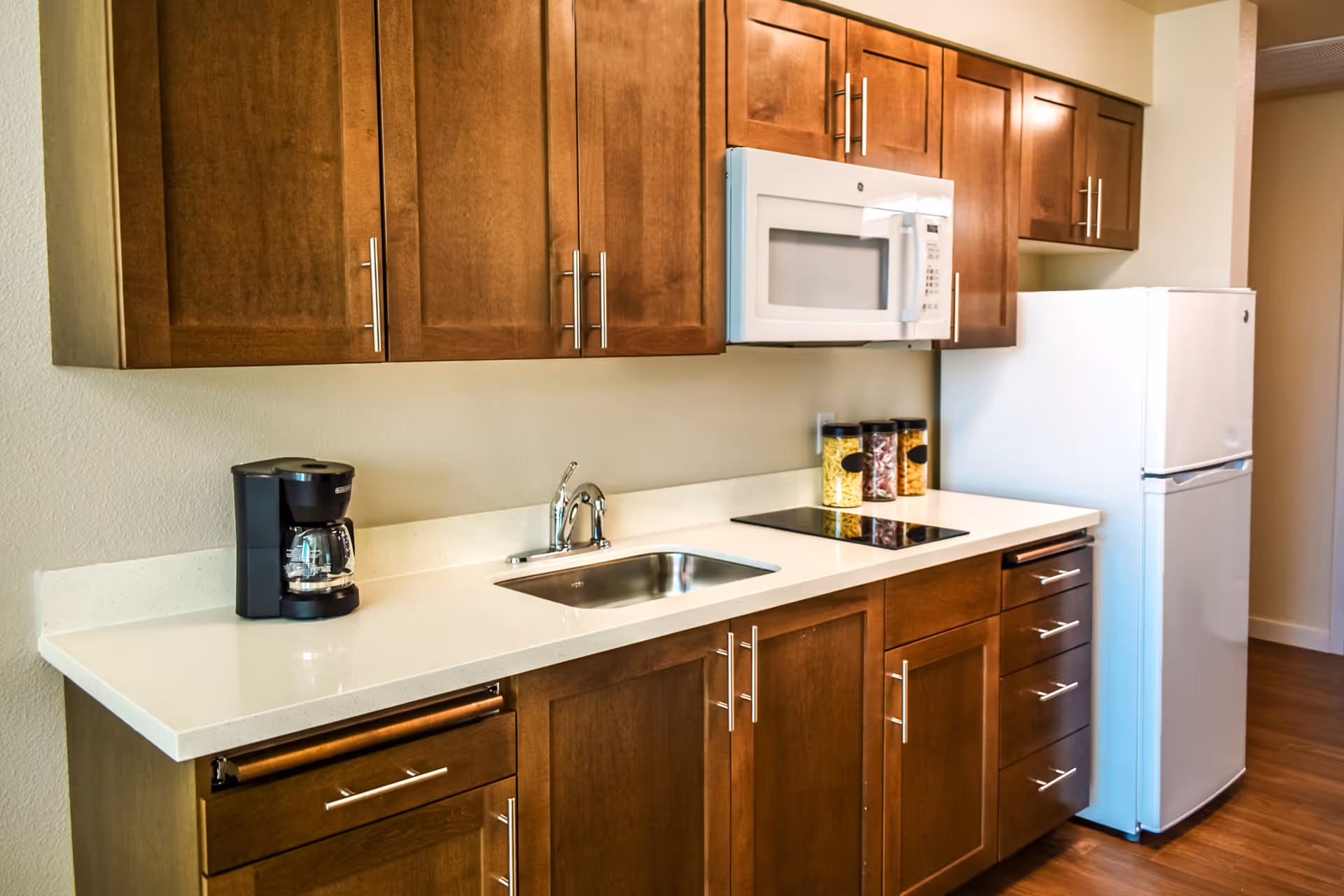 A modern kitchen with wooden cabinets, a white countertop, a stainless steel sink, a black coffee maker, a white microwave, a white refrigerator, and three jars filled with pasta and other dry goods.