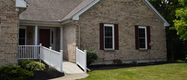 Brick senior living home exterior with a front porch, white railings, and trimmed lawn.