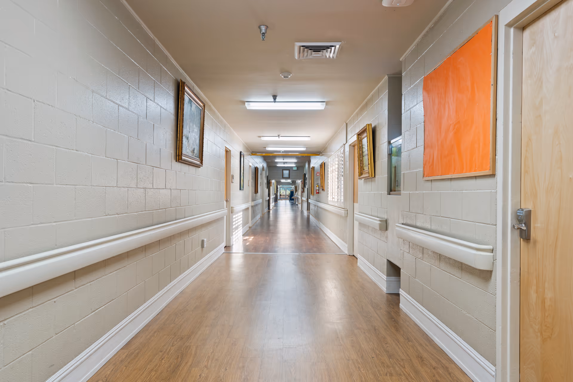 A long, well-lit hallway in a senior living facility with beige walls, wooden doors, handrails along both sides, framed paintings on the walls, and a wooden floor.