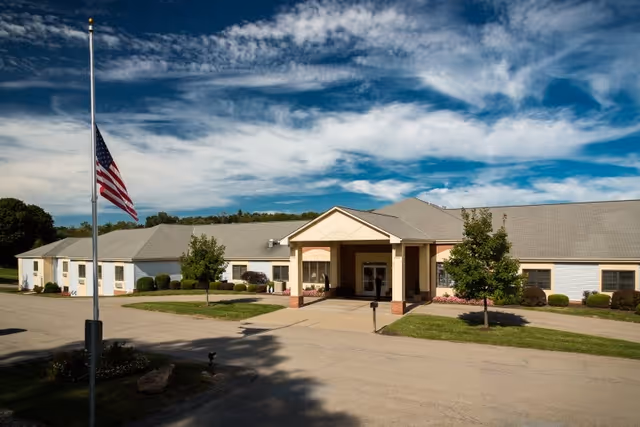 Exterior view of a single-story senior living facility building with a covered entrance, surrounded by small trees and bushes under a partly cloudy blue sky. An American flag is flying at half-mast on a flagpole in front of the building.