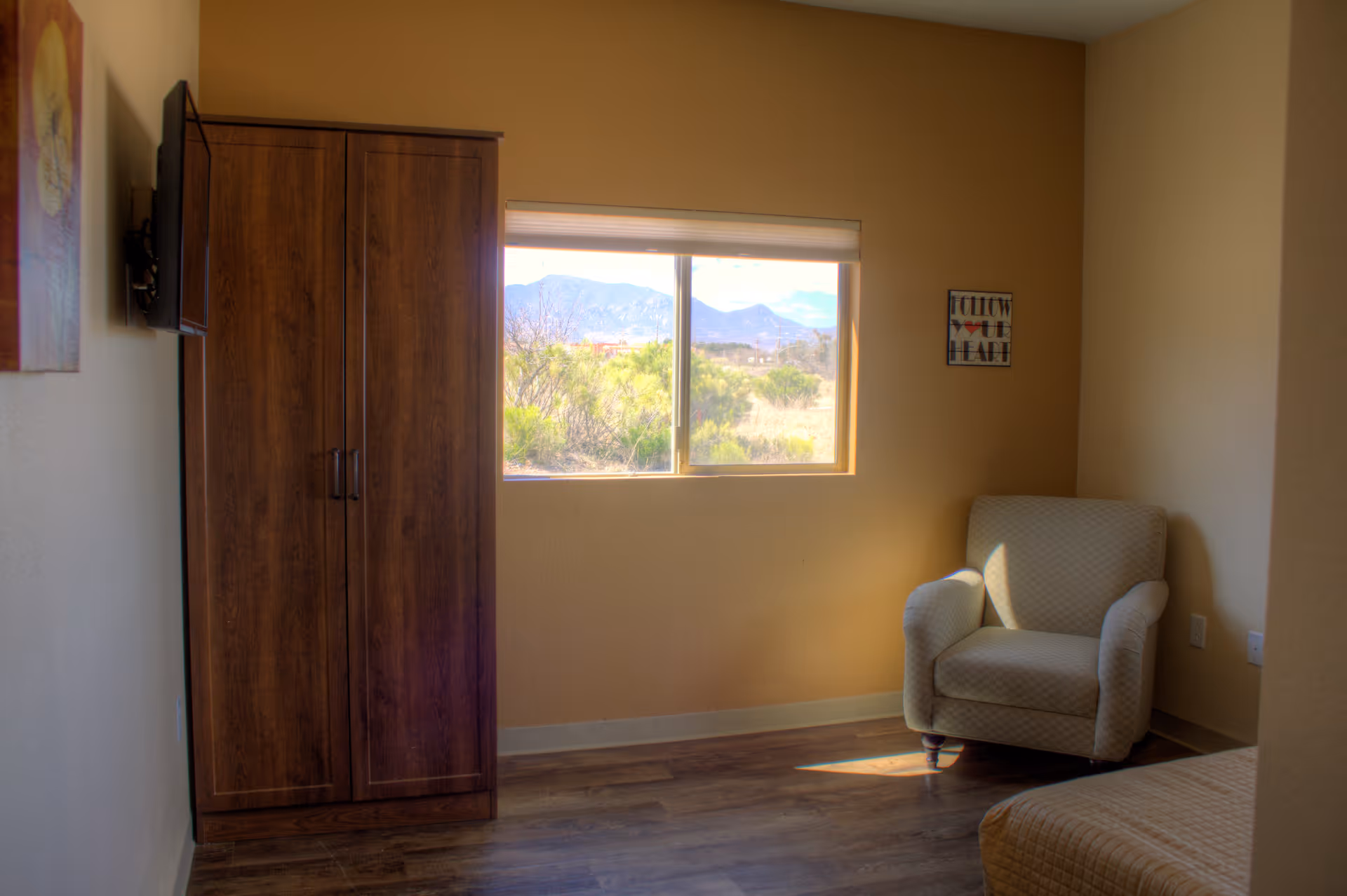 A cozy room with a wooden wardrobe, a wall-mounted TV, a comfortable armchair, and a window showing a scenic view of mountains and greenery outside. The walls are painted beige, and there is a small framed artwork on the wall.