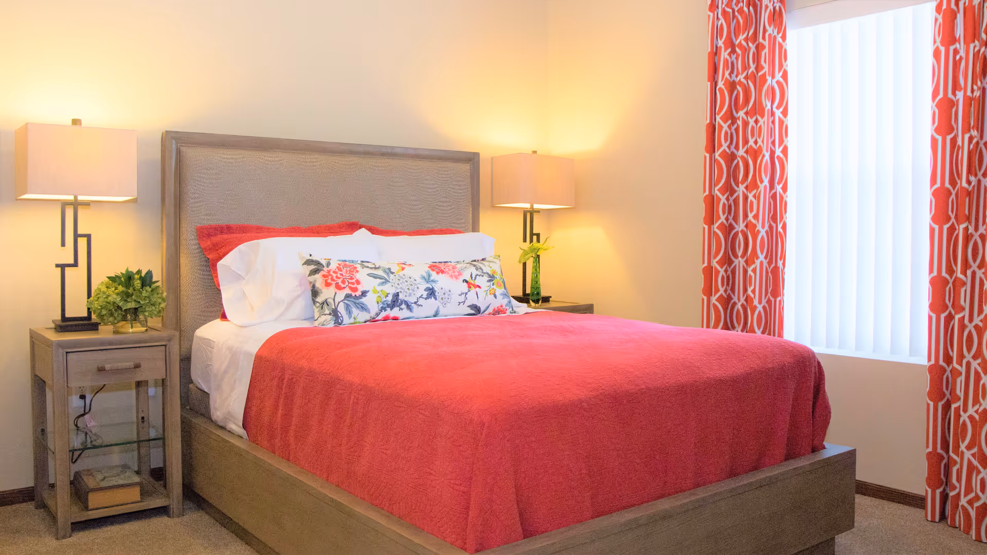 A bedroom with a wooden bed frame and a red bedspread. The bed has white pillows and a decorative floral pillow. There are two wooden nightstands on either side of the bed, each with a modern lamp and a small plant. The room has beige walls and a window with red and white patterned curtains.