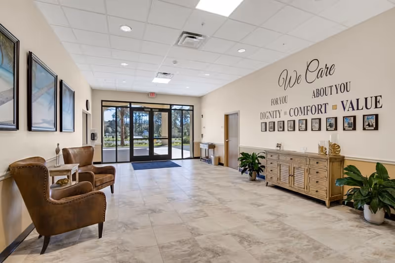 A spacious and well-lit lobby area with large glass doors leading outside. The room features two brown leather armchairs with a small table between them on the left side, and a wooden sideboard with decorative items and framed photos on the right wall. Above the sideboard, there is a wall decal that reads 'We Care For You About You Dignity Comfort Value'. There are also two potted plants on the floor near the sideboard and a few framed pictures on the left wall.