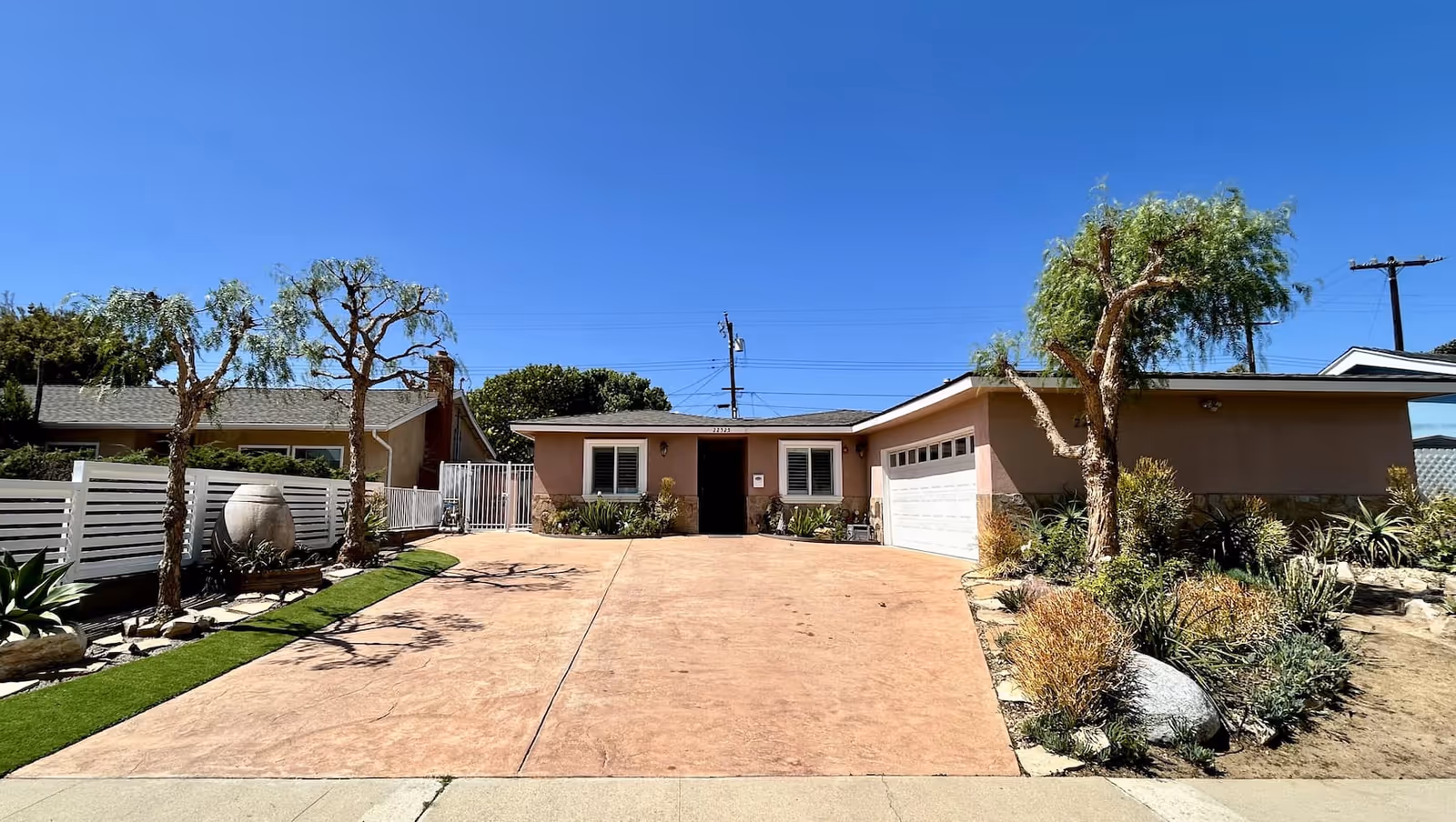Front exterior view of a single-story house with a wide driveway, two small trees, and landscaped garden beds on either side. The house has a light brown exterior with a white garage door and two windows flanking the front entrance. The sky is clear and blue.