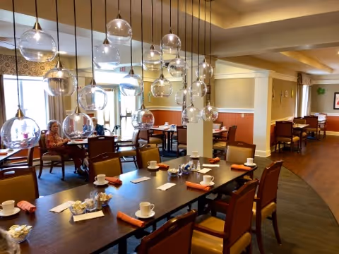 Dining room with a long table set with cups and napkins, hanging glass globe pendant lights, and people seated near windows.