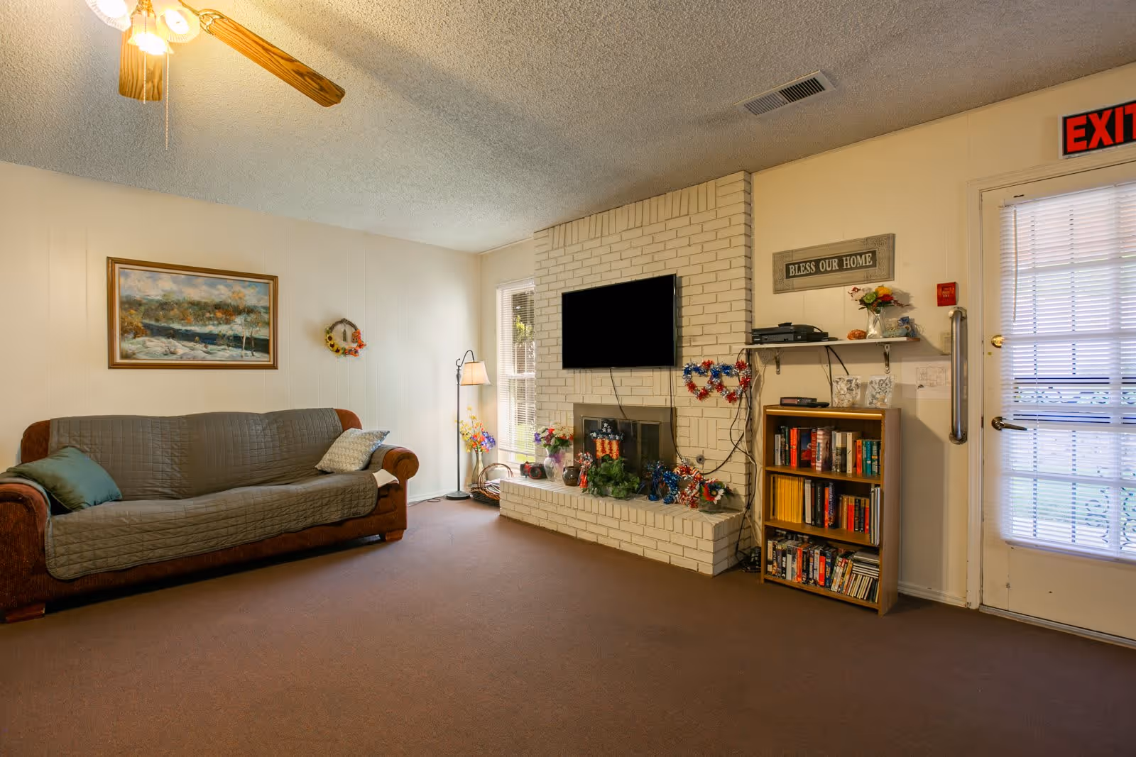 A cozy living room with a brown couch covered by a gray quilted cover, a floor lamp, a white brick fireplace with a mounted flat-screen TV above it, and a small bookshelf filled with books and DVDs. The room has beige walls, a ceiling fan with lights, a painting on the wall, and a door with a window covered by blinds. There is a sign above the bookshelf that reads 'BLESS OUR HOME' and some decorative items on the fireplace and shelf.