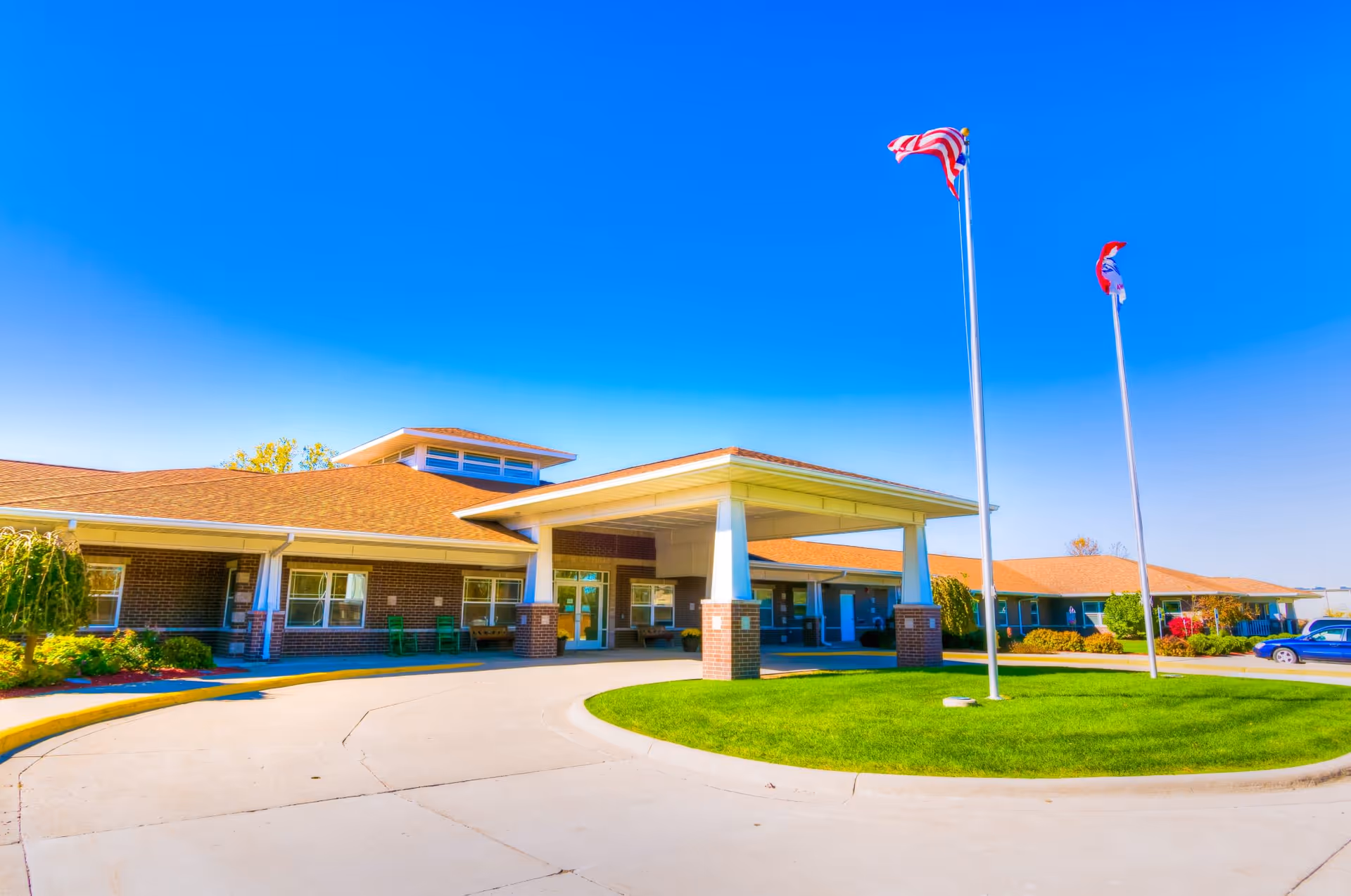 Exterior view of River Valley Place of Ottumwa, a single-story brick building with a covered entrance supported by white columns. Two flagpoles with flags are positioned on a grassy circular island in front of the entrance. The sky is clear and blue.