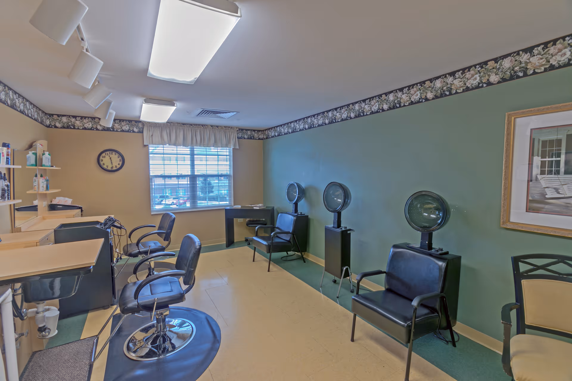 Interior view of a hair salon area in a senior living facility with salon chairs, hair dryers, a window with blinds, a clock on the wall, and shelves with hair products.