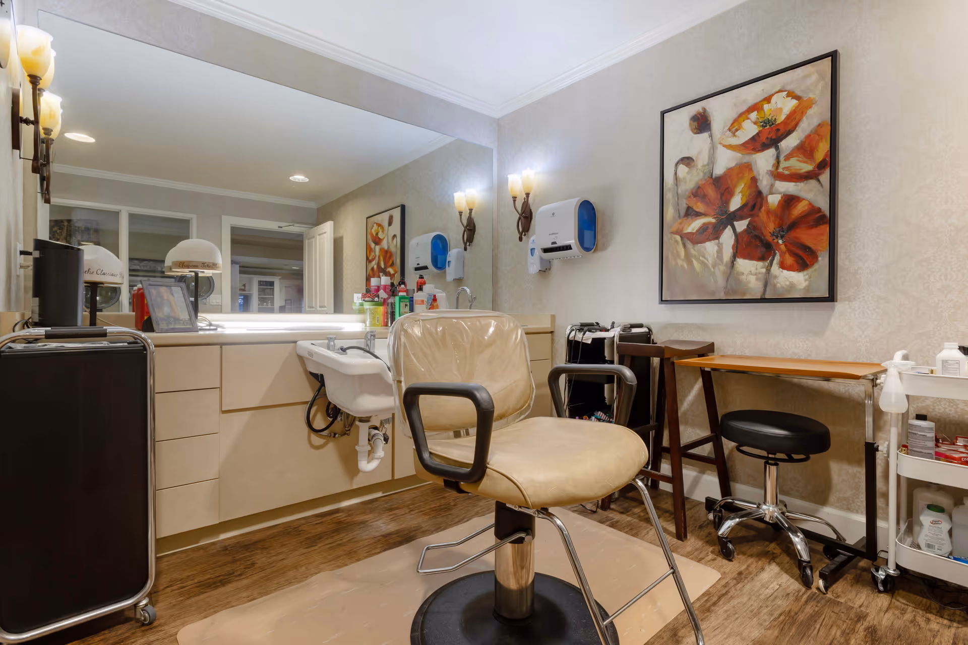 Interior of a hair salon area in a senior living facility with a beige salon chair in front of a large mirror, a white sink for washing hair, various hair care products on the counter, a black rolling cart, a small wooden table with a black rolling stool, and a colorful painting of red and orange flowers on the wall.