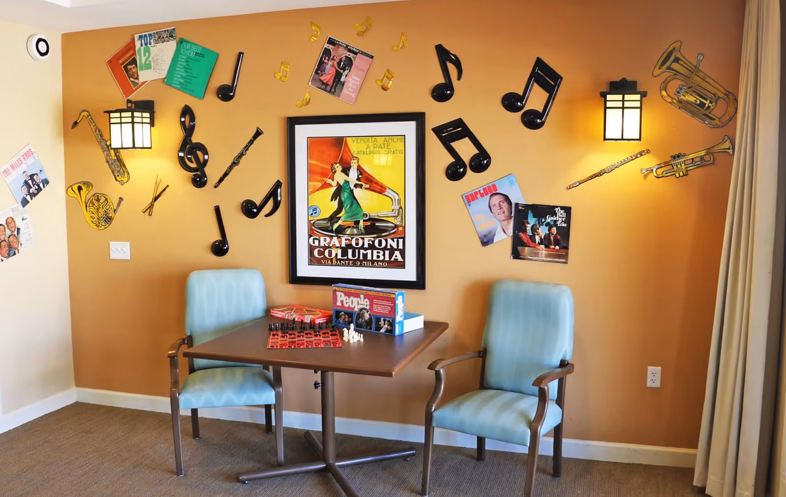 A cozy corner in a memory care facility with two light blue cushioned chairs around a small wooden table. The table has a chessboard and some books on it. The wall behind is decorated with musical notes, instruments, and vintage music album covers, along with a framed vintage poster. Two wall-mounted lamps provide warm lighting.