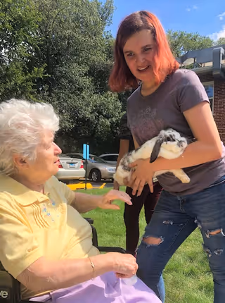 An elderly woman sitting in a wheelchair outdoors reaching out to pet a white and brown rabbit held by a younger woman with red hair wearing a purple t-shirt and ripped jeans. Trees, parked cars, and a building are visible in the background under a blue sky.