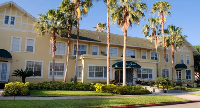Exterior view of a two-story senior living facility building with beige walls, multiple windows, green awnings, and several tall palm trees in front. The building is surrounded by well-maintained green lawns and shrubs under a clear blue sky.