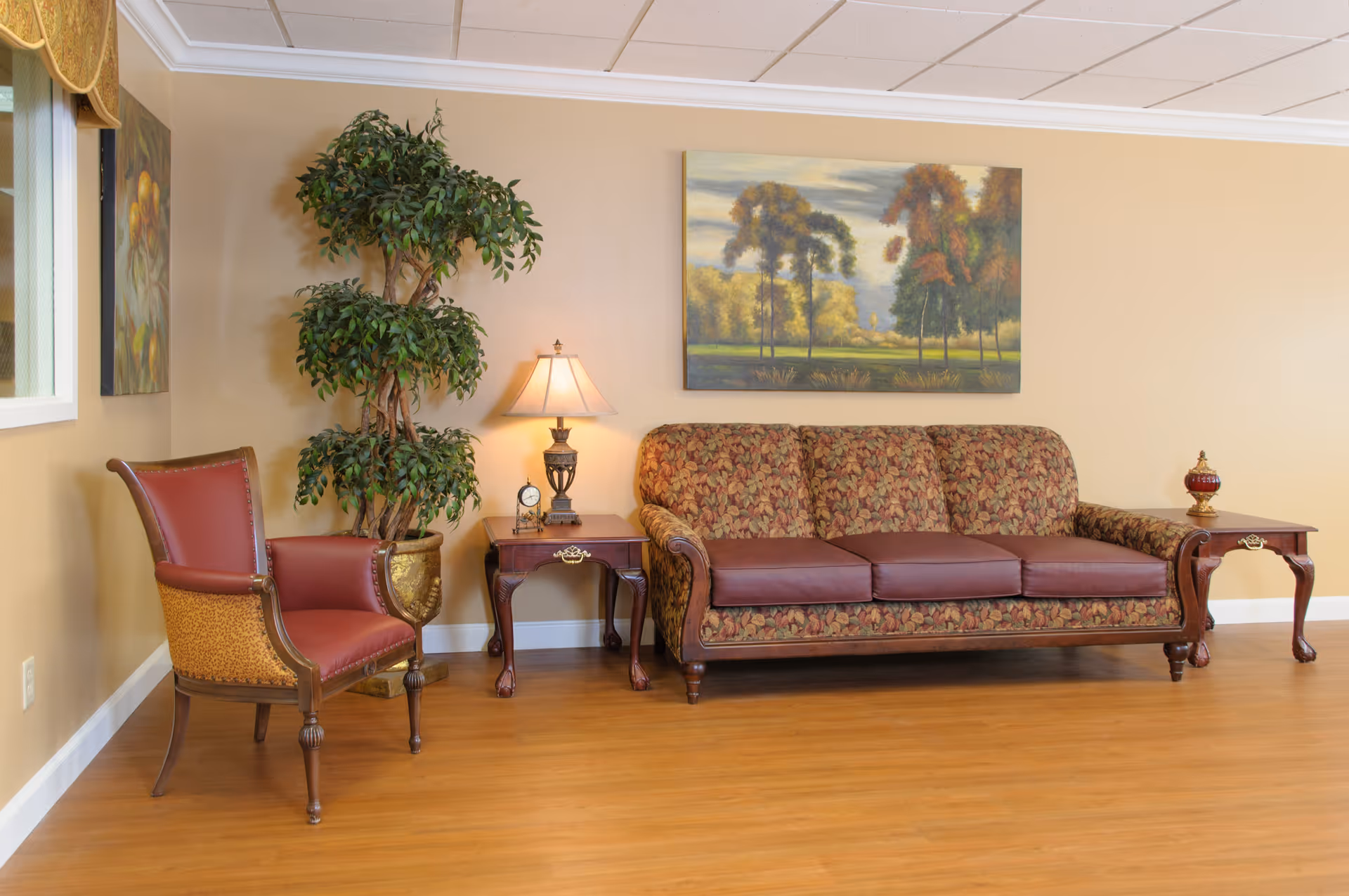 A furnished sitting area with a patterned three-seat sofa, red leather armchair, side tables with a lamp and decor, a potted plant, and a landscape painting on a beige wall.