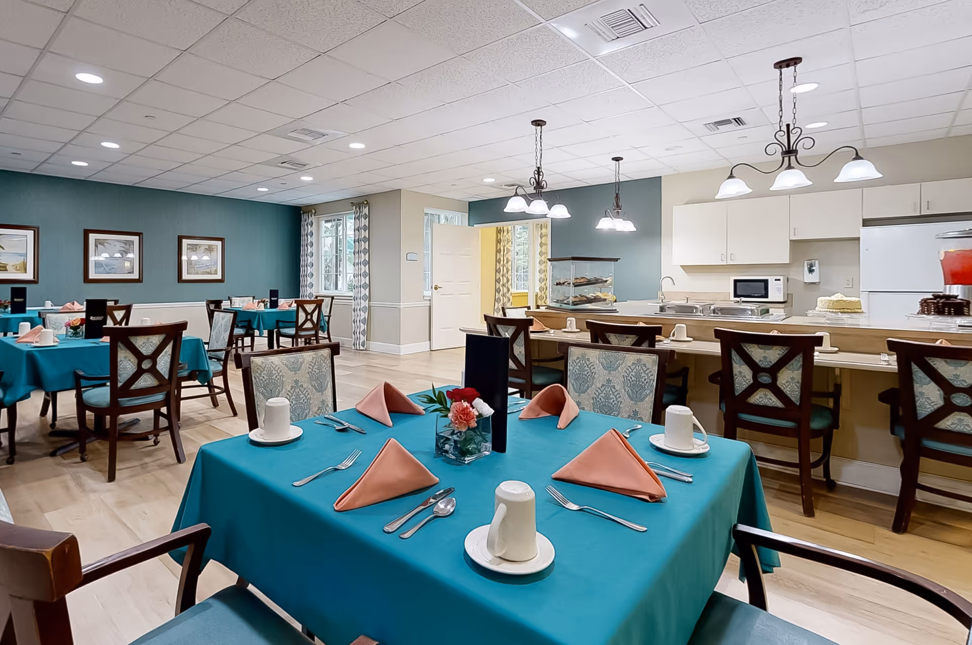 A dining room in a senior living facility with tables covered in teal tablecloths, set with pink folded napkins, white cups, and silverware. The room has wooden chairs with patterned cushions, a kitchen area with white cabinets, a microwave, a refrigerator, and pendant lights hanging from the ceiling. There are framed pictures on the teal-colored wall and windows with patterned curtains.