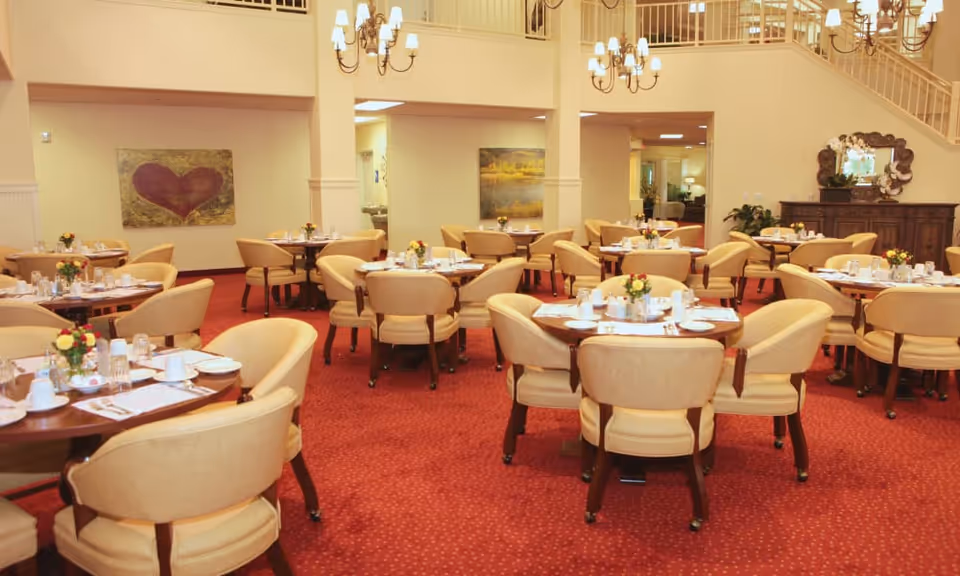 A spacious dining room with multiple round tables set with plates, cups, and silverware. Each table has beige upholstered chairs and small floral centerpieces. The room features red carpet, cream-colored walls, chandeliers, and artwork on the walls. There is a staircase and a wooden sideboard with a mirror in the background.