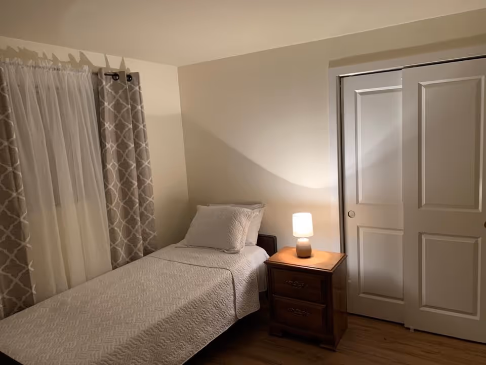 A small bedroom with a single bed covered in a white quilt and a matching pillow. Next to the bed is a wooden nightstand with two drawers, on top of which sits a small lamp emitting warm light. The room has light-colored walls, a window with sheer white curtains and patterned gray drapes, and a closet with white sliding doors.