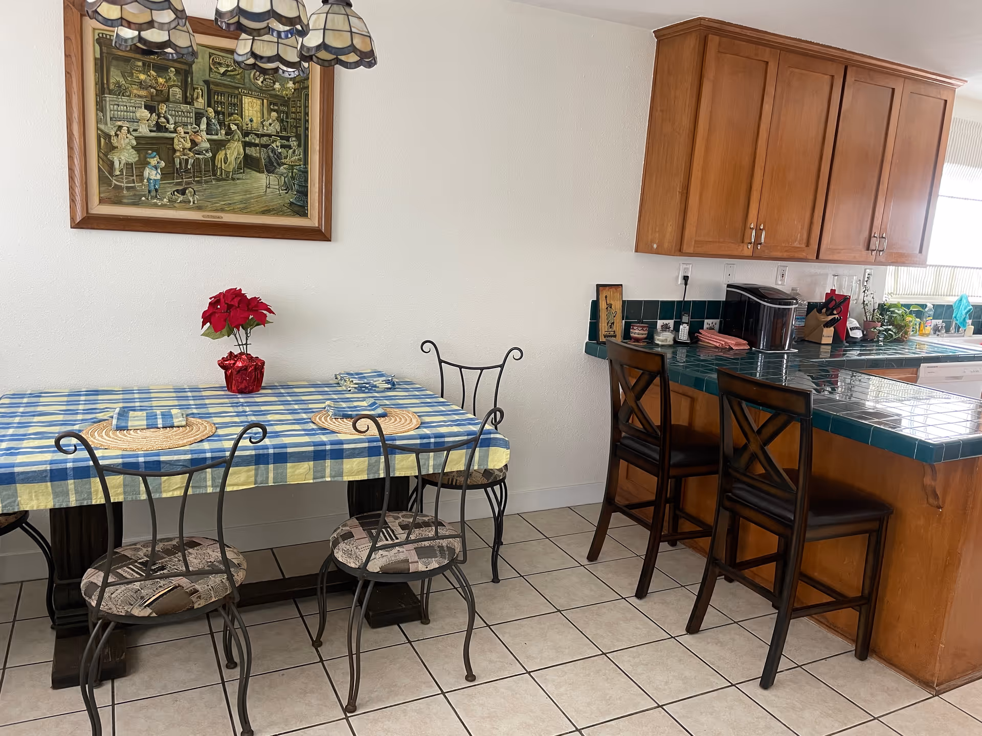 A dining area with a table covered by a blue and yellow checkered tablecloth, set with placemats and napkins. Four metal chairs with patterned cushions surround the table. To the right, there is a kitchen counter with green tiled countertops and two wooden bar stools. Above the counter are wooden cabinets. A framed painting depicting a bar scene hangs on the wall above the dining table. The floor is tiled in a light color.