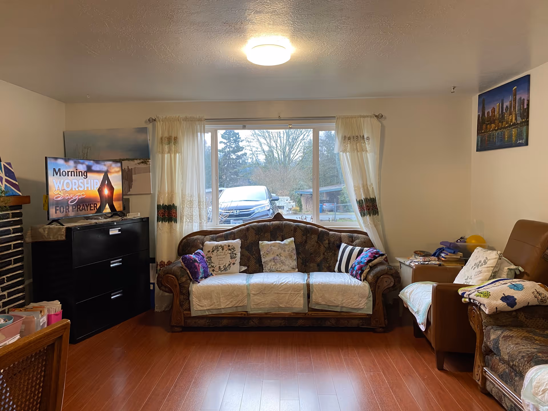 A cozy living room with a large window covered by white curtains with floral embroidery. In front of the window is a patterned sofa with several decorative pillows and white covers. To the right is a brown armchair with a white cover and a side table holding books and other items. On the left side, there is a black cabinet with a TV on top displaying the text 'Morning Worship Songs for Prayer'. The room has wooden flooring and light-colored walls with a cityscape painting on the right wall.