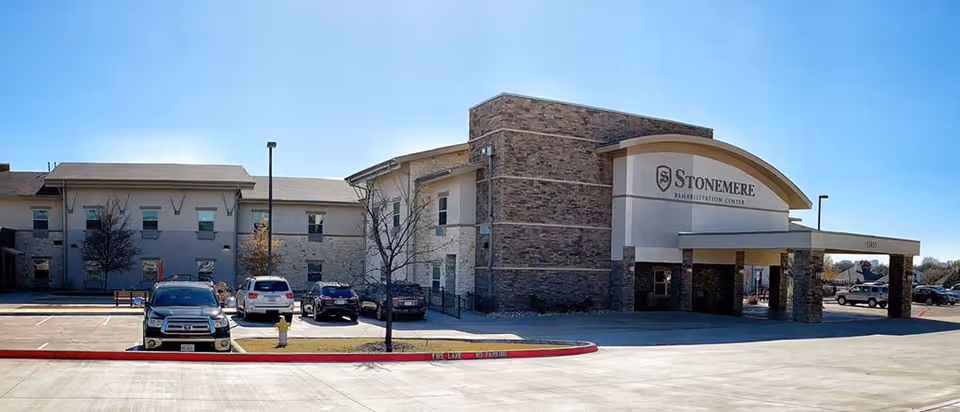 Exterior view of Stonemere Rehabilitation Center building with a parking lot in front. Several cars are parked, and the building has a combination of stone and light-colored walls under a clear blue sky.
