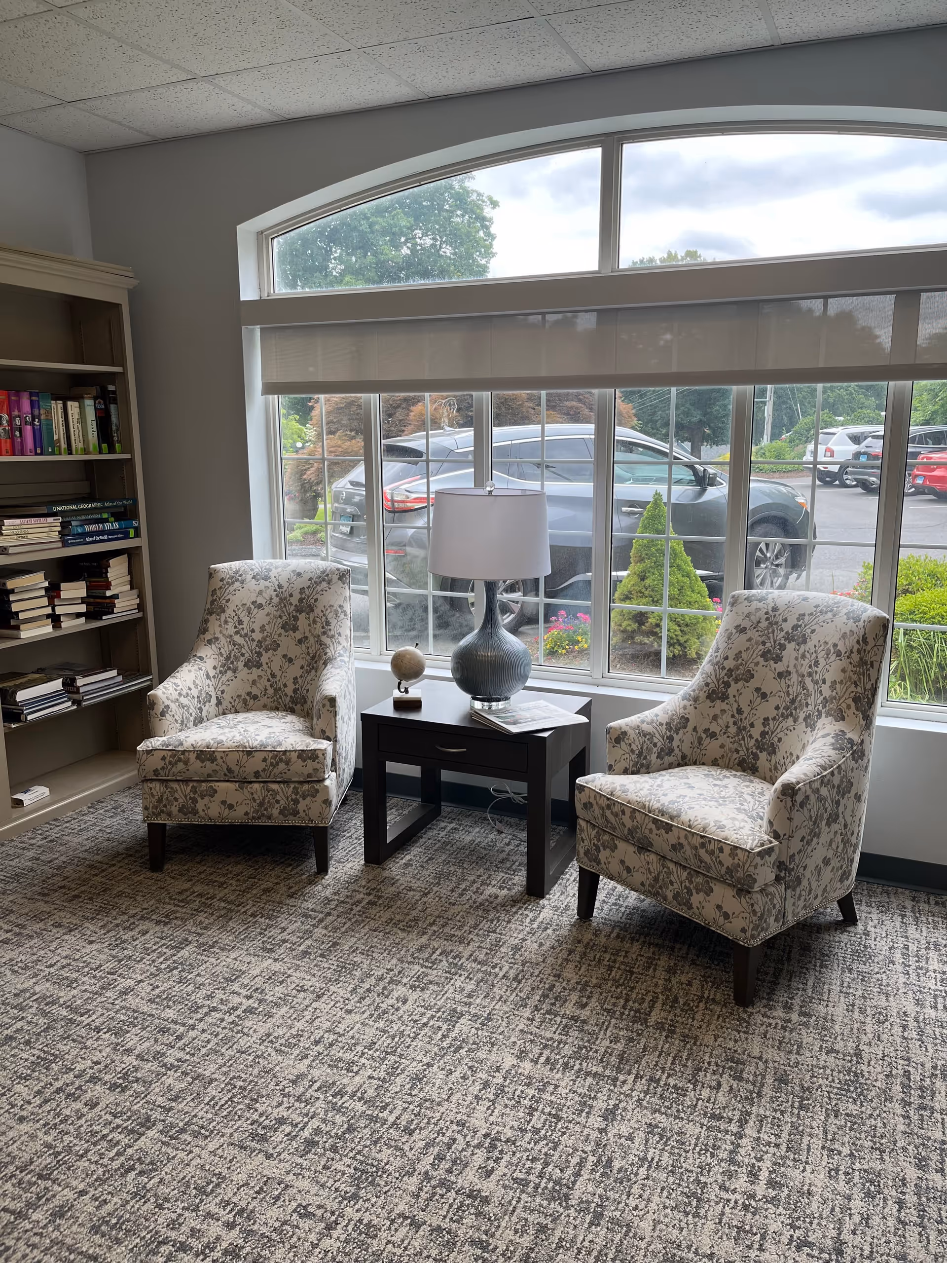 Two patterned armchairs flank a small side table with a lamp in a carpeted seating area by a large window overlooking a parking lot.