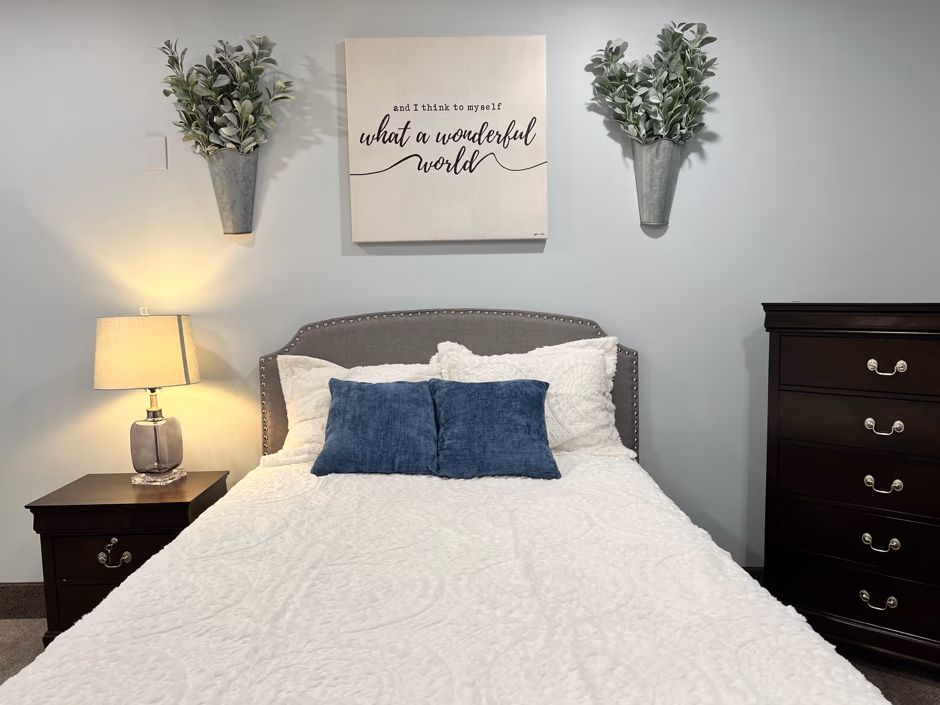 A neatly made bed with a gray upholstered headboard, white textured bedding, and two blue pillows. On the left side of the bed is a dark wooden nightstand with a lamp emitting warm light. Above the bed is a wall decoration with the text 'and I think to myself what a wonderful world' flanked by two wall-mounted metal planters holding green leafy plants. To the right of the bed is a dark wooden dresser with silver handles.