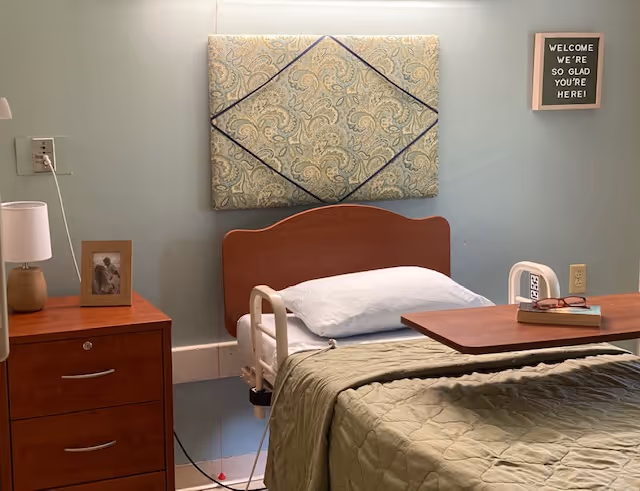 A senior living facility bedroom with a hospital-style bed covered with a green quilt. There is a wooden headboard and a padded decorative panel on the wall above it. A wooden nightstand with a lamp and a framed photo is next to the bed. On the wall, a small sign reads 'WELCOME WE'RE SO GLAD YOU'RE HERE!'. A wooden overbed table holds a pair of glasses and a book.
