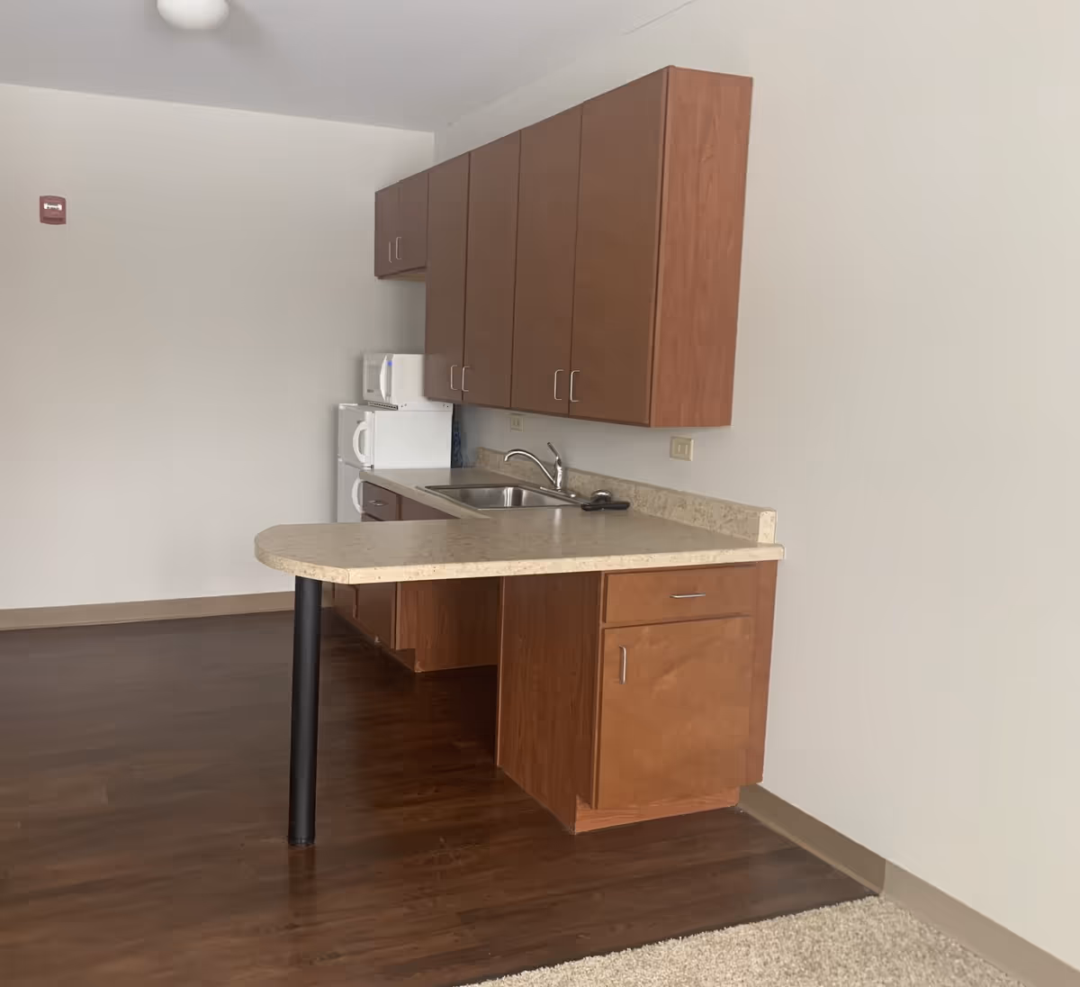 A small kitchen area with wooden cabinets, a beige countertop with a rounded edge supported by a black leg, a stainless steel sink with a faucet, a white microwave, and a white refrigerator. The floor is dark wood, and the walls are plain white.