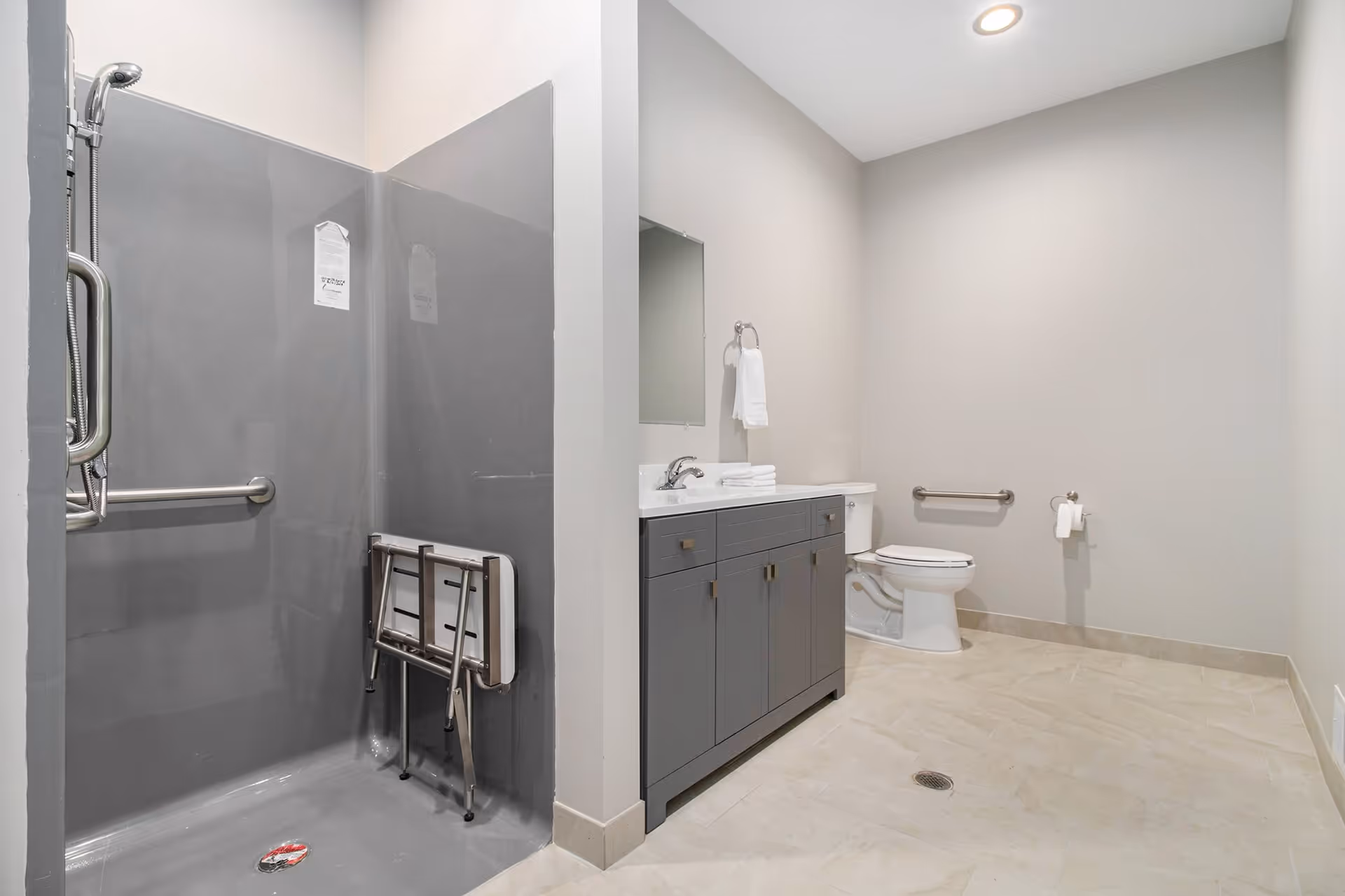 A spacious bathroom with a gray walk-in shower equipped with a foldable seat and grab bars, a gray vanity with a sink and mirror, a toilet with a grab bar, and beige tiled floor.