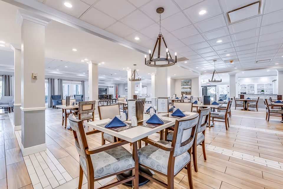 Bright, spacious senior living dining room with multiple set tables and chairs, chandeliers, and large windows.